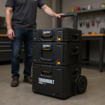 Take the product from the reference image (preserve its exact shape, colours, branding, and orientation). Place it naturally in this setting at a realistic scale, with no people in the frame: photorealistic photo of a worker standing beside a three-tier modular tool cart with stackable boxes in black and gray, positioned in a professional workshop or garage, industrial workbench in background, clear bright lighting highlighting the cart's wheeled base and modular design, organized tools visible, professional environment, sharp focus